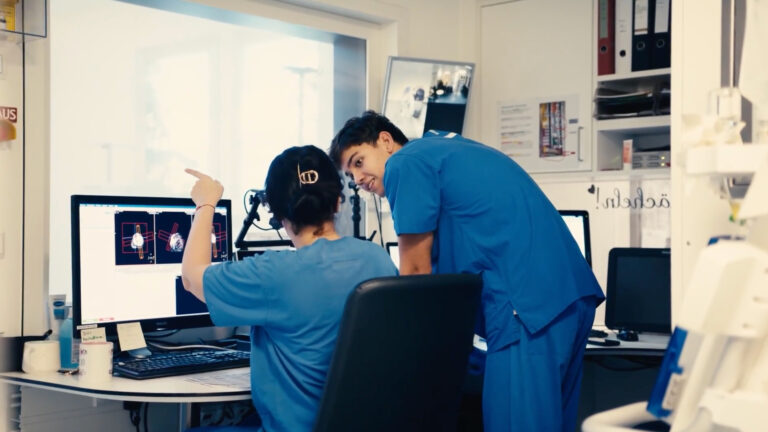 Zwei Fachkräfte der Radiologietechnologie in blauer Arbeitskleidung analysieren medizinische Schnittbilder am Computerbildschirm im Kontrollraum eines Landeskrankenhauses Bregenz.