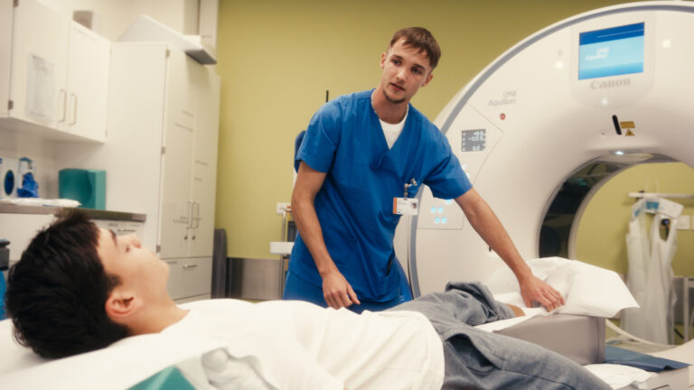 Ein medizinischer Mitarbeiter in blauer Kleidung betreut einen Patienten auf der Liege eines Computertomographen.