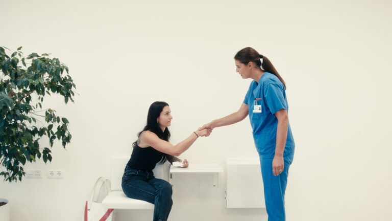 Eine Pflegekraft in blauer Arbeitskleidung gibt einer auf einer Bank sitzenden Frau die Hand.