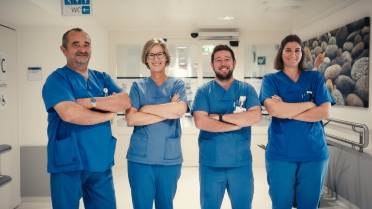 Vier lächelnde Mitarbeiter in blauer Arbeitskleidung stehen mit verschränkten Armen nebeneinander auf einem Krankenhausflur.