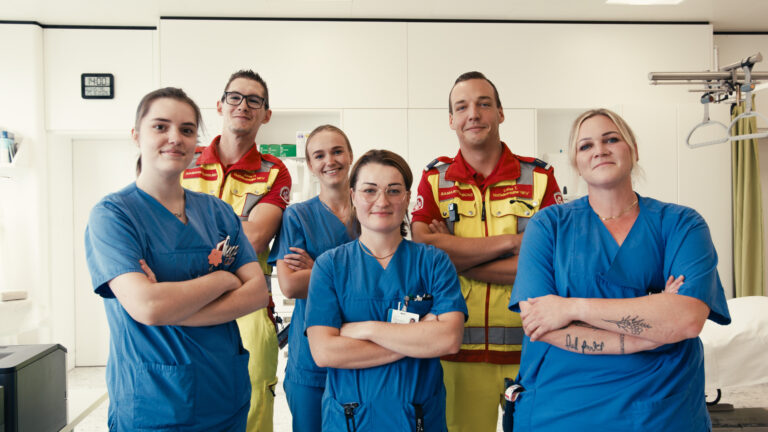 Vier Pflegekräfte und zwei Personen aus dem Rettungsdienst stehen lächelnd in einem Behandlungsraum.