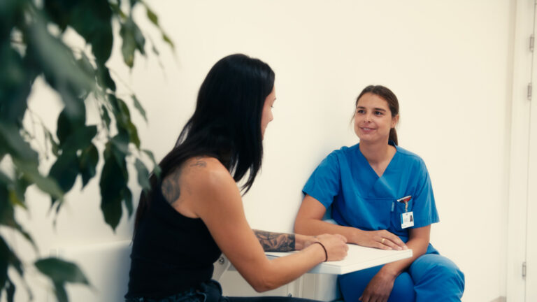 Eine lächelnde Pflegekraft in blauer Kleidung sitzt einer Frau an einem Tisch gegenüber und führt ein Gespräch mit ihr.