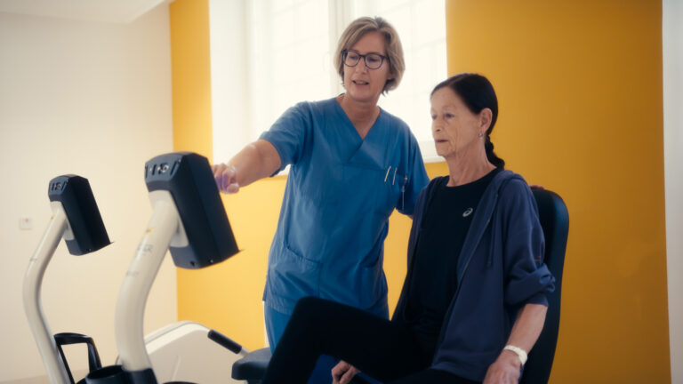 Eine Pflegekraft erklärt einer älteren Patientin auf einem Bewegungstrainer die Einstellungen am Monitor.