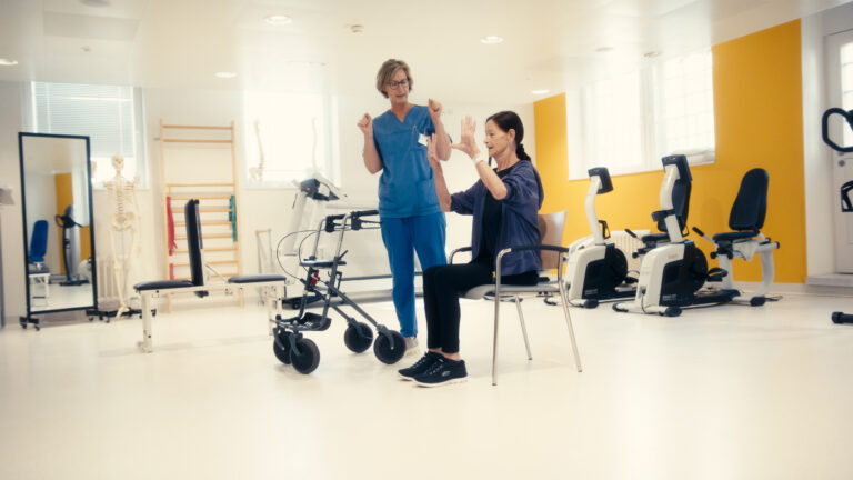 Eine Pflegekraft leitet eine ältere Patientin auf einem Stuhl bei Bewegungsübungen an.