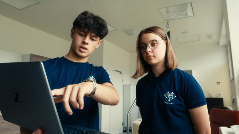 Zwei Mitarbeiter:innen aus der IT-Abteilung im dunkelblauen Teamshirt arbeiten gemeinsam an einem Laptop.