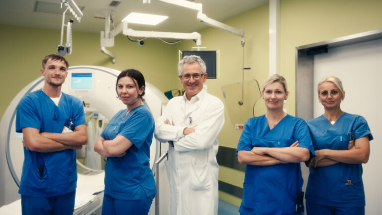 Fünfköpfiges Radiologie-Team, bestehend aus einem Arzt im weißen Kittel und vier Radiologietechnologinnen und -technologen in blauer Dienstkleidung, mit verschränkten Armen vor einem modernen Computertomographen (CT).