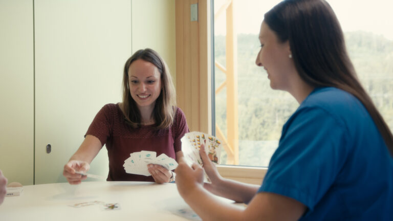 Eine Patientin und eine Pflegekraft spielen gemeinsam Karten an einem Tisch.