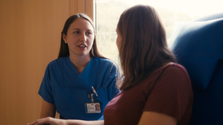 Eine Pflegekraft in blauer Kleidung spricht aufmerksam mit einer Patientin.
