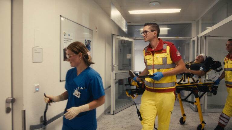 Rettungskräfte und eine Pflegekraft bringen einen Patienten auf einer Fahrtrage durch den Krankenhausflur.