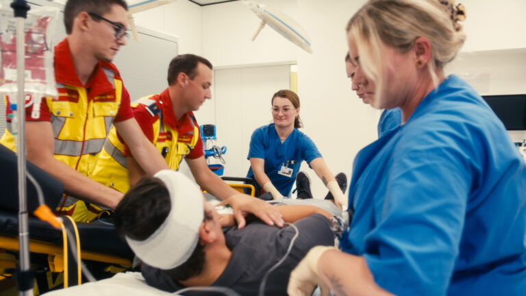 Ein Team aus Rettungsdienst und Pflegekräften lagert einen Patienten mit Kopfverband gemeinsam auf eine Behandlungsliege um.