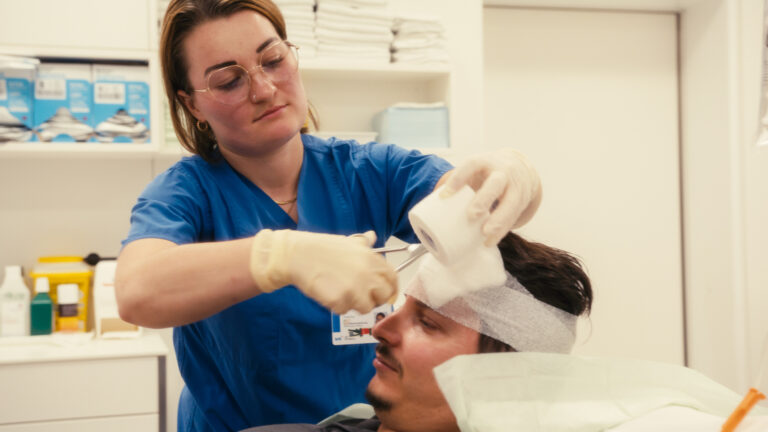 Eine Pflegekraft schneidet den Kopfverband eines Patienten im Behandlungsraum zurecht.