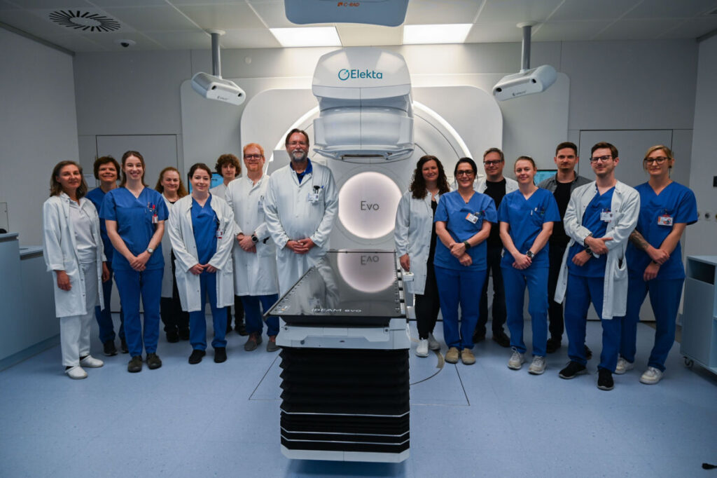 Ein großes medizinisches Team in weißer und blauer Arbeitskleidung steht gemeinsam vor einem modernen Linearbeschleuniger für die Strahlentherapie im Behandlungsraum.
