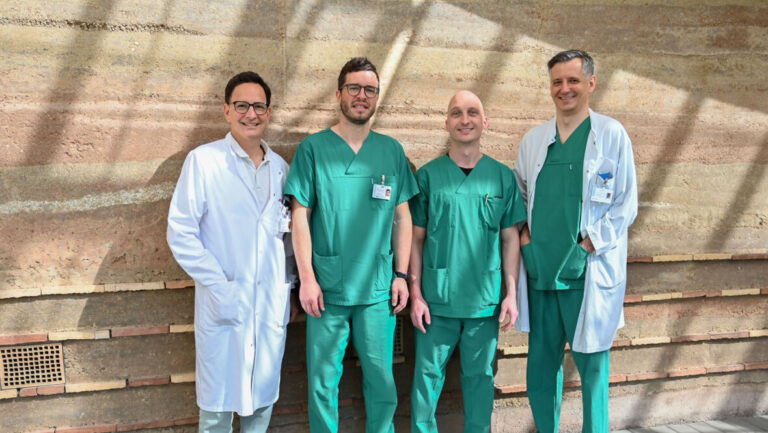 Vier Mediziner in grünen OP-Kasacks und weißen Arztkitteln stehen nebeneinander im Flur.