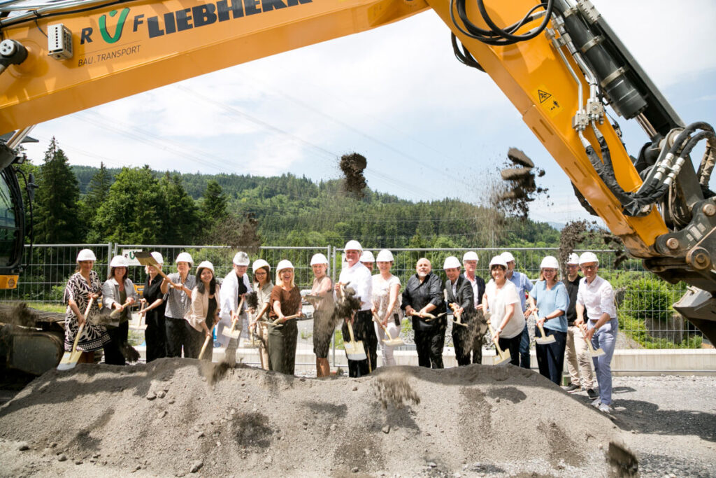 Eine große Gruppe von Beteiligten mit weißen Bauhelmen und Spaten steht nebeneinander aufgereiht auf der Baustelle vor einem Bagger.