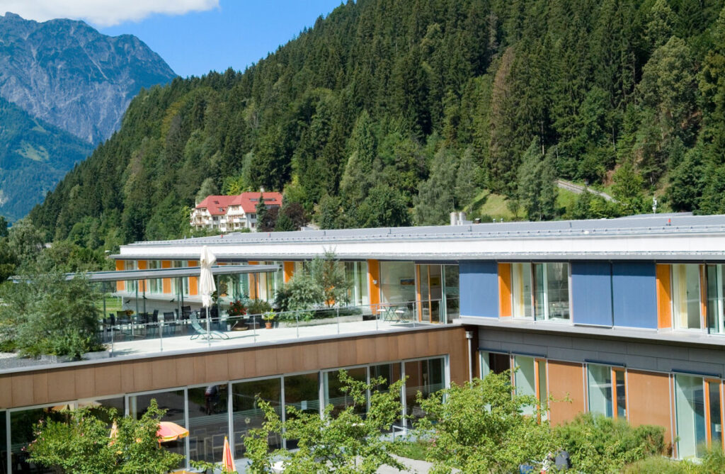 Ein modernes Gebäude mit großen Fensterfronten und Sonnenterrasse liegt direkt vor einer dicht bewaldeten Bergkulisse.