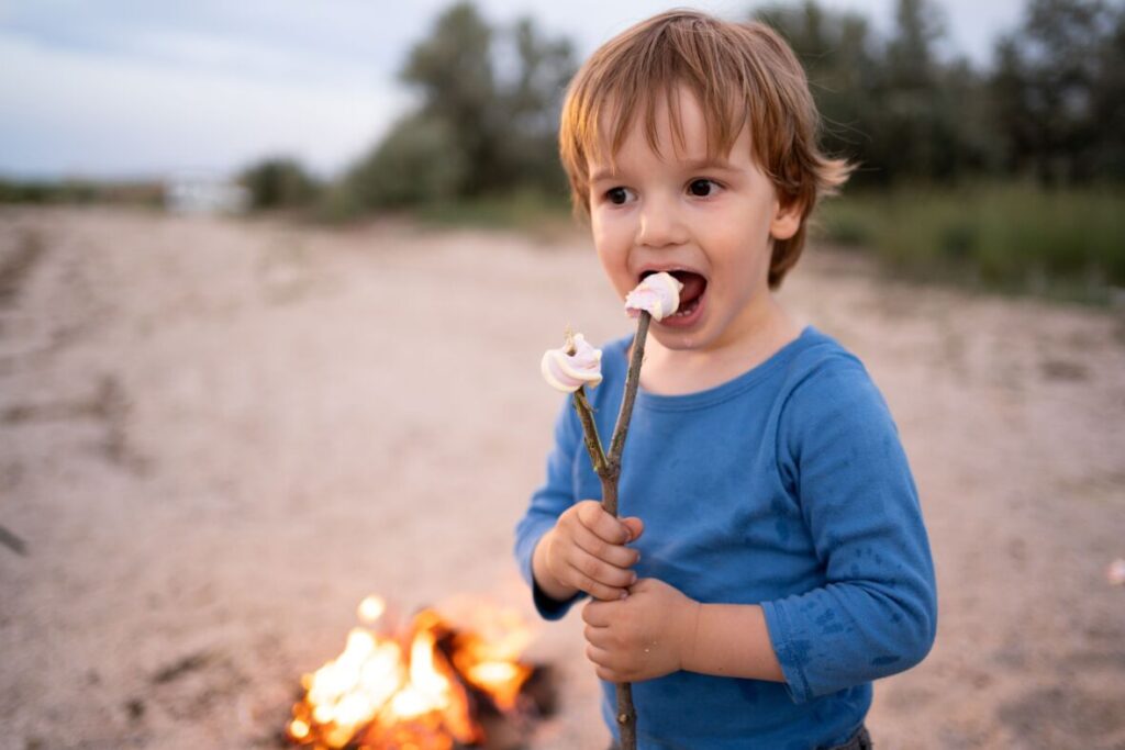Ein kleiner Junge im blauen Oberteil isst draußen vor einem Lagerfeuer einen Marshmallow von einem Holzstock.