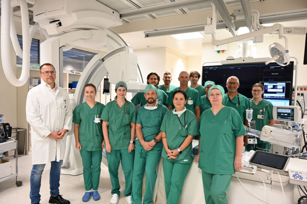 Ein medizinisches Team in grüner OP-Kleidung und ein Arzt im weißen Kittel stehen gemeinsam vor der Behandlungsanlage im Herzkatheterlabor.
