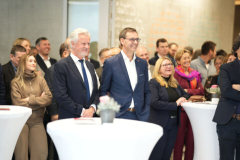 Aufmerksame Gäste in eleganter Kleidung verfolgen das Eröffnungsprogramm an Stehtischen.