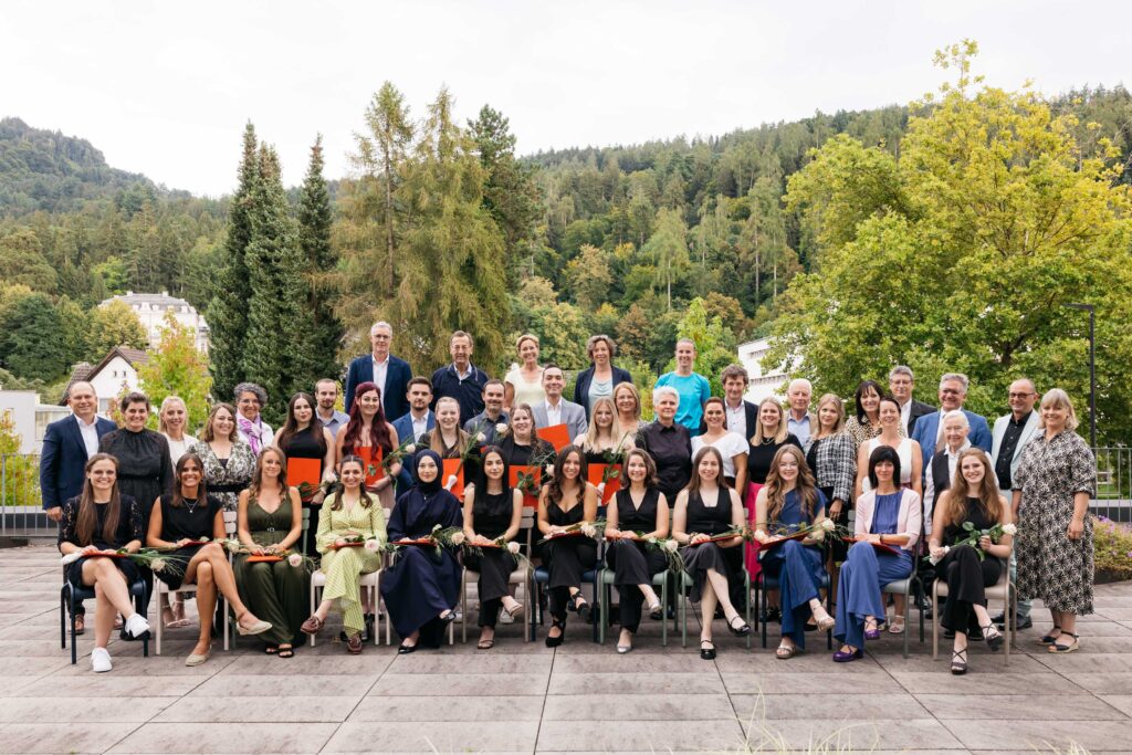 Große Gruppe von Absolventinnen und Absolventen der diplomierten Gesundheits- und Krankenpflege (DGKP) mit roten Diplommappen und Rosen im Freien vor bewaldeter Kulisse in Bregenz.
