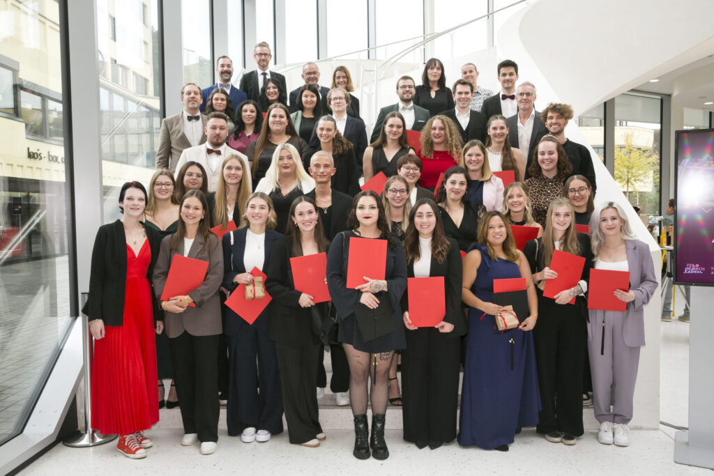 Absolventengruppe der diplomierten Gesundheits- und Krankenpflege hält stolz ihre roten Diplommappen auf einer modernen Treppe im Foyer in Feldkirch.