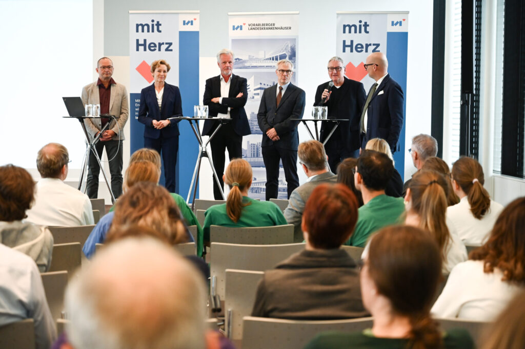 Podiumsdiskussion im Rahmen einer Veranstaltung der Vorarlberger Landeskrankenhäuser (LKH) vor Publikum. Fünf Referenten stehen an Stehtischen, im Hintergrund hängen Banner mit dem Logo der LKH und dem Slogan "mit Herz".