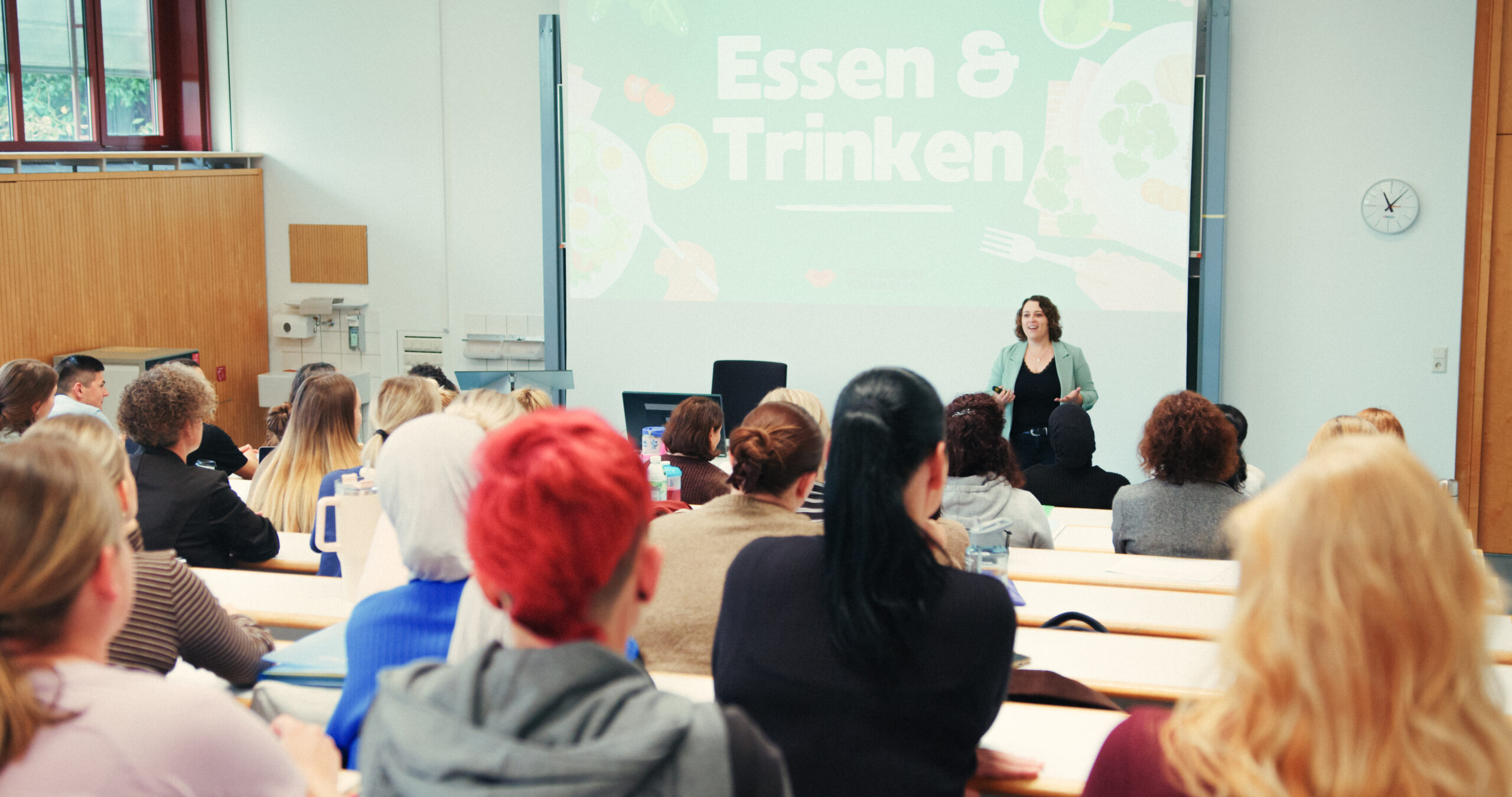 Blick aus dem Publikum auf eine Dozentin vor einer Präsentation zum Thema "Essen & Trinken" im Hörsaal der Pflegeschule Vorarlberg.